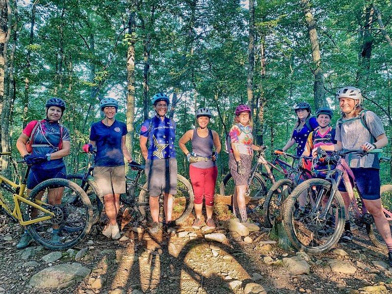 Several women with mountain bikes and wearing helmets smiling at the camera around sunset